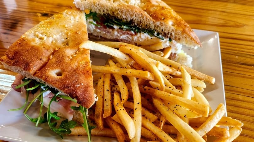 Focaccia sandwich with prosciutto, arugula, and cheese, served with a side of french fries.