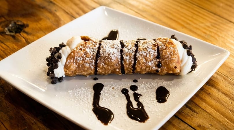 A cannoli dessert, drizzled with chocolate syrup and powdered sugar, on a white rectangular plate.