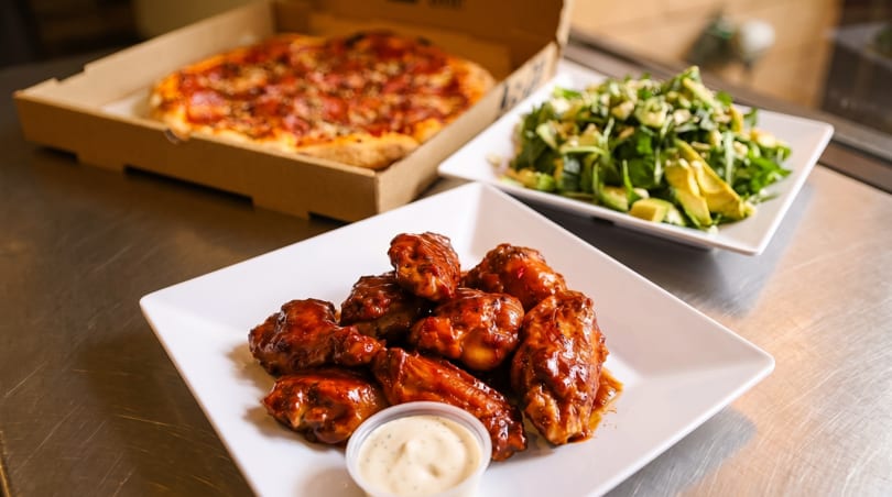 A pizza in a box, a plate of chicken wings with dipping sauce, and a green salad.